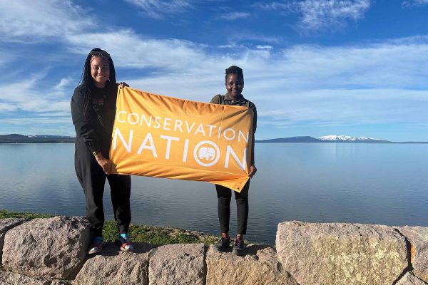 two Black women holding an orange flag that reads conservation nation, while standing on a rock structure with water and mountains in the background