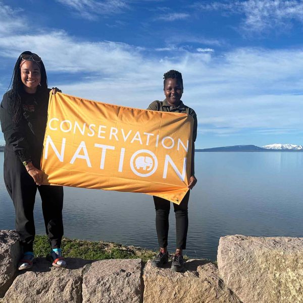 two Black women holding an orange flag that reads conservation nation, while standing on a rock structure with water and mountains in the background