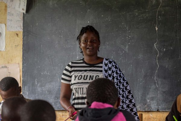 a black woman wearing black and white patterned clothing standing in front of a chalkboard while addressing students