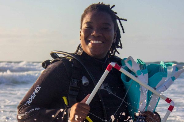 a Black woman wearing scuba diving gear smiling and walking in the surf with crashing waves in the background